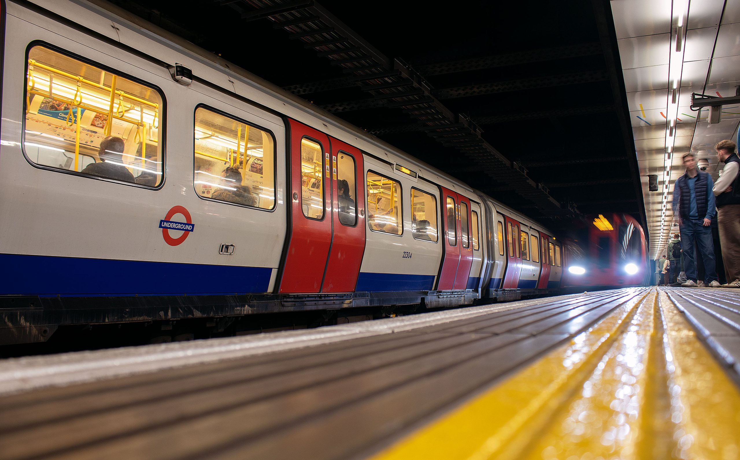 London_Underground_train_stopped_at_a_Tube_station_platform_with_passengers_waiting_and_another_train_approaching_in_the_background